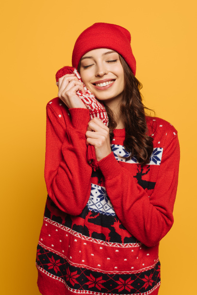 happy girl in hat and red ornamental sweater holding mittens near face with closed eyes isolated on yellow - Foto, Imagen