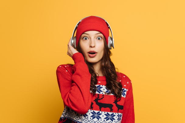 zaskoczona dziewczyna w kapeluszu, słuchawki bezprzewodowe i czerwony sweter ozdobny patrząc na aparat odizolowany na żółto - Zdjęcie, obraz