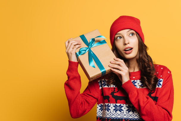 curious girl in hat and red ornamental sweater holding gift box on yellow background - Фото, изображение