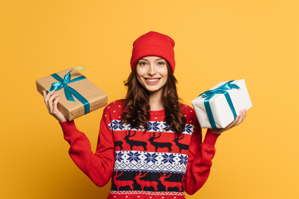 happy girl in hat and red ornamental sweater holding gift boxes on yellow background - Photo, Image
