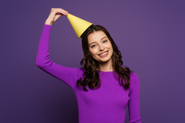 happy girl touching party cap while smiling at camera on purple background - Foto, Bild