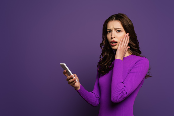 шокована дівчина торкається обличчя, тримаючи смартфон на фіолетовому фоні
 - Фото, зображення