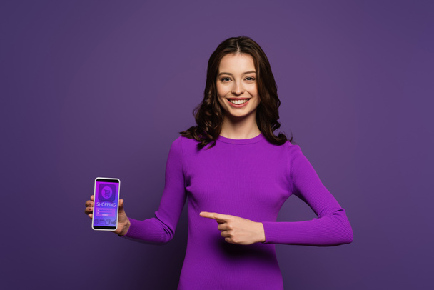 chica sonriente apuntando con el dedo en el teléfono inteligente con aplicación de compras en la pantalla sobre fondo púrpura
 - Foto, imagen