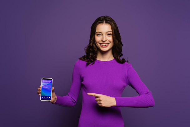mosolygós lány ujjal mutatva okostelefon foglalási alkalmazás a képernyőn lila háttér - Fotó, kép