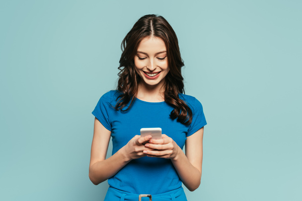 cheerful girl smiling while chatting on smartphone isolated on blue - Photo, Image