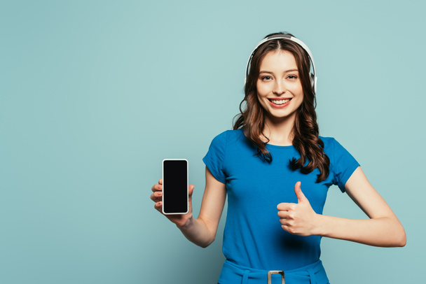 cheerful girl in wireless headphones showing thumb up while holding smartphone with bank screen on blue background - Фото, зображення