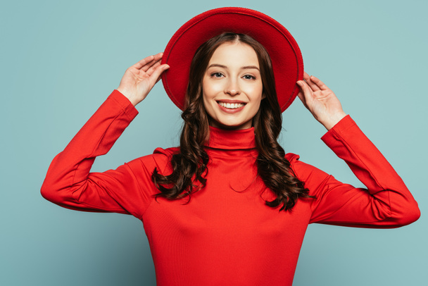 smiling stylish girl touching brims of hat while looking at camera on blue background - Foto, imagen