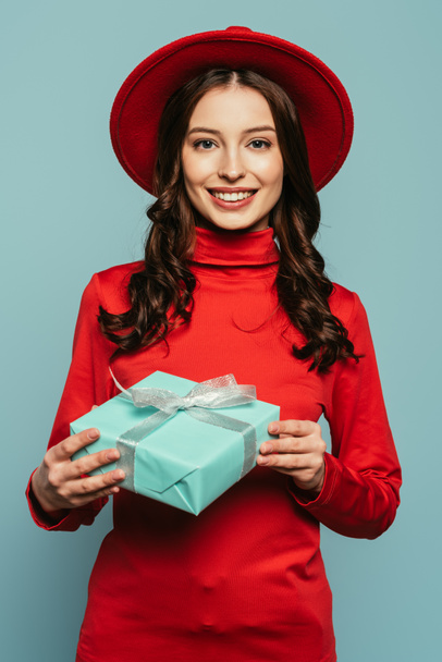 cheerful stylish girl smiling at camera while holding gift box isolated on blue - Foto, imagen