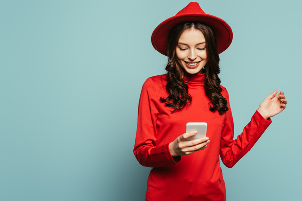 cheerful stylish girl smiling while chatting on smartphone on blue background - Photo, Image