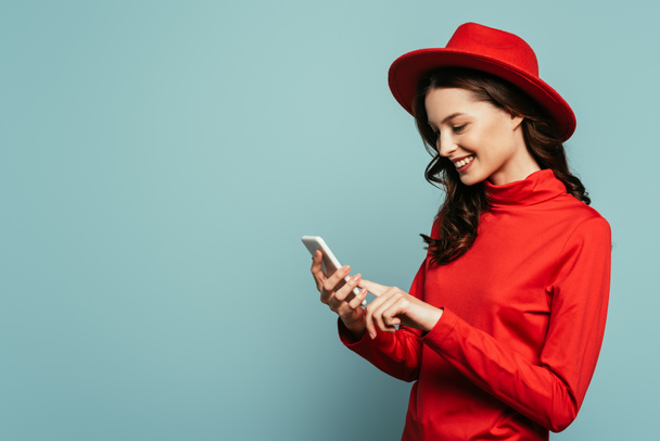 happy stylish girl smiling while chatting on smartphone isolated on blue - Photo, Image