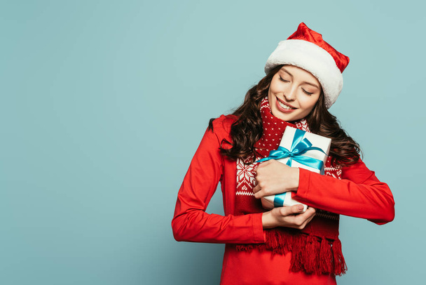 happy girl in santa hat hugging gift box with closed eyes on blue background - Fotoğraf, Görsel
