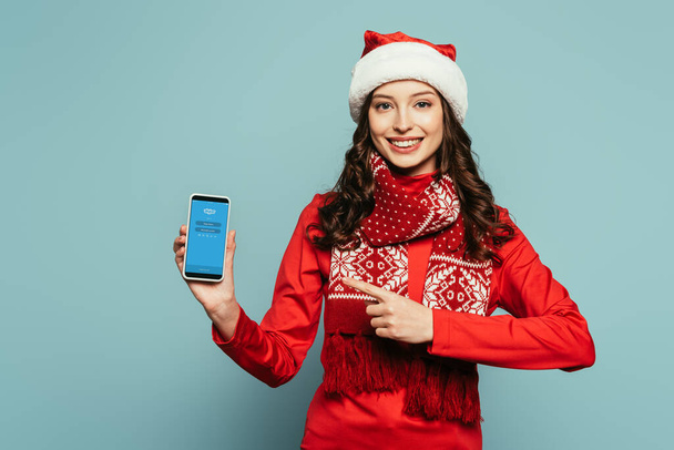 1KYIV, UKRAINE - NOVEMBER 29, 2019: happy girl in santa hat and red sweater pointing with finger at smartphone with Skype app on screen on blue background - Fotografie, Obrázek