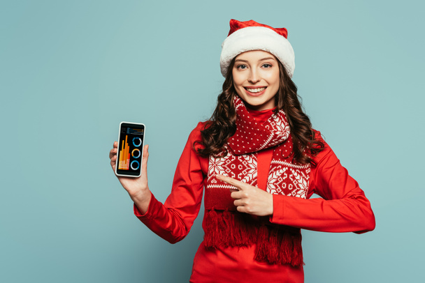 happy girl in santa hat and red sweater pointing with finger at smartphone with graphs and charts on screen on blue background - Photo, Image