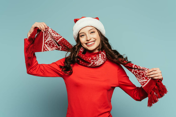 happy girl in santa hat touching red scarf and smiling at camera on blue background - Photo, Image