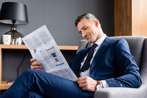 hombre de negocios sonriente en traje sentado en sillón y leyendo el periódico con letras de negocios
  - Foto, Imagen
