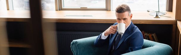 tiro panorâmico de homem de negócios em terno beber café e sentado em poltrona
  - Foto, Imagem