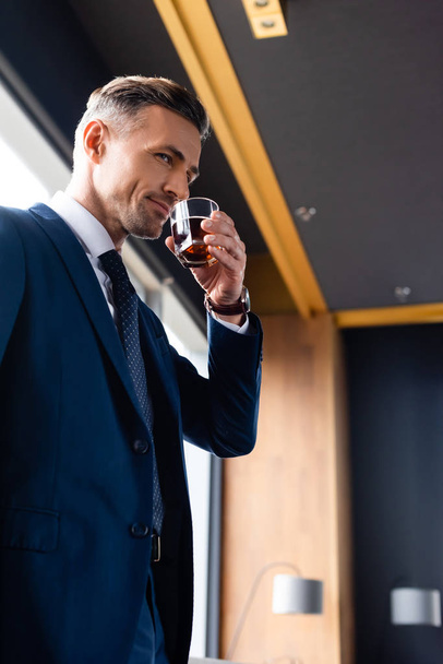 low angle view of smiling businessman in suit drinking cognac - Fotó, kép