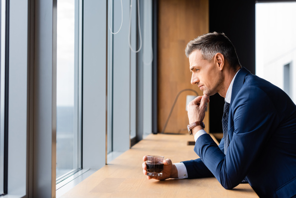 side view of pensive businessman in suit holding glass and looking through window  - Photo, image