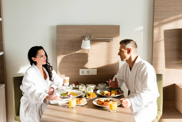 zijaanzicht van vriend en vriendin in badjassen praten en ontbijten - Foto, afbeelding