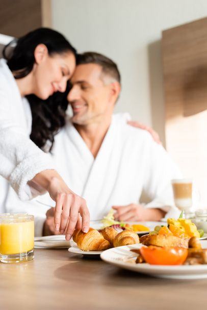 valikoiva painopiste hymyilevä tyttöystävä kylpytakki halaus poikaystävä ja ottaen croissant hotellissa
  - Valokuva, kuva