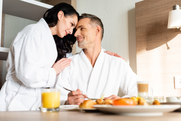 selective focus of smiling girlfriend in bathrobe hugging boyfriend during breakfast  - Fotografie, Obrázek