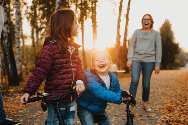 Mãe caucasiana alegre ensinando seus filhos sorridentes a andar de bicicleta durante uma noite colorida de outono
 - Foto, Imagem