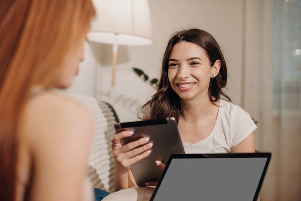 Brunette caucasian girl with a tablet is looking at her red haired friend who is using a laptop sitting on a sofa - Foto, imagen