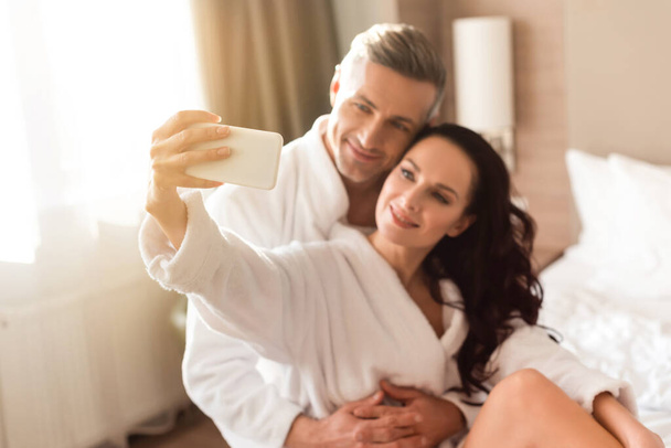 selective focus of boyfriend hugging smiling girlfriend in bathrobe and she taking selfie in hotel - 写真・画像