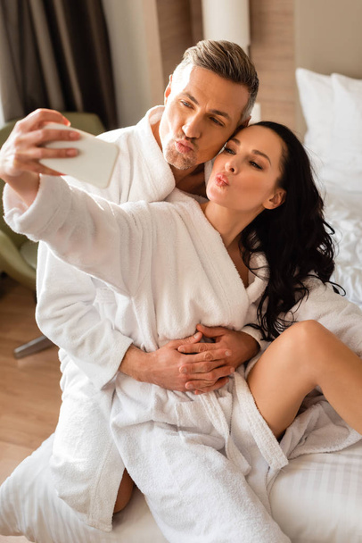 boyfriend hugging girlfriend in bathrobe and she taking selfie and showing kiss in hotel - Photo, Image