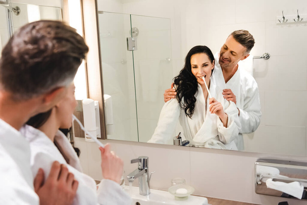 selective focus of boyfriend hugging smiling girlfriend and she brushing teeth in hotel - Fotó, kép