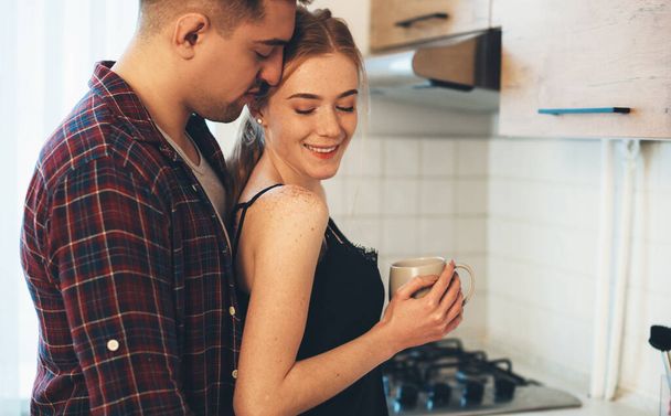 Vorsichtiger Mann umarmt sein kaukasisches Mädchen mit Sommersprossen und roten Haaren, während er in der Küche einen Kaffee trinkt - Foto, Bild