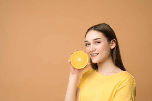 Beautiful brunette with healthy skin holds half of orange near her face and looking at the camera, isolated with copyspace - Foto, afbeelding