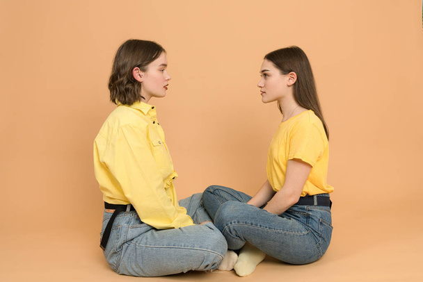 Two attractive girls in blue jeans and yellow shirts sittig against each other and looking into each others eyes, isolated on orange background - Photo, Image