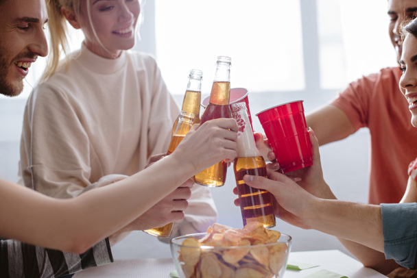 cropped view of happy friends clinking bottles of beer and plastic cups with soda  - 写真・画像