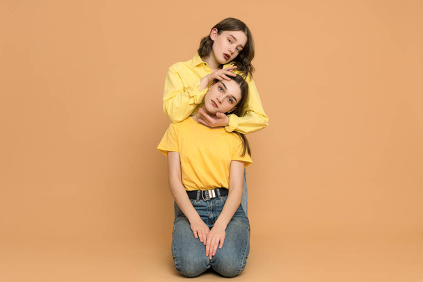 Pretty ladies in yellow shirts having fun on floor in orange studio, brunette woman holds her hands on her sisters head, both looking at the camera - Foto, Imagen