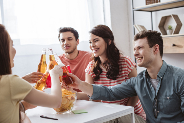 šťastní přátelé cinkání s lahvemi a poháry při hraní názvu hry doma - Fotografie, Obrázek