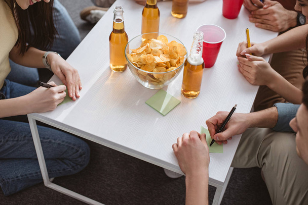 abgeschnittene Ansicht von Freunden, die auf klebrige Zettel schreiben, während sie am Tisch ein Namensspiel mit Getränken und Chips spielen - Foto, Bild
