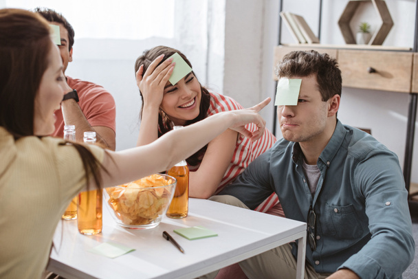 selective focus of girl pointing with hand at man with sticky note on forehead while playing name game with friends - Foto, Imagen