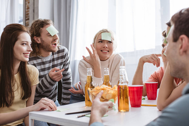 fröhliche Freunde mit klebrigen Zetteln auf der Stirn beim Namensspiel am Tisch mit Bier und Chips - Foto, Bild