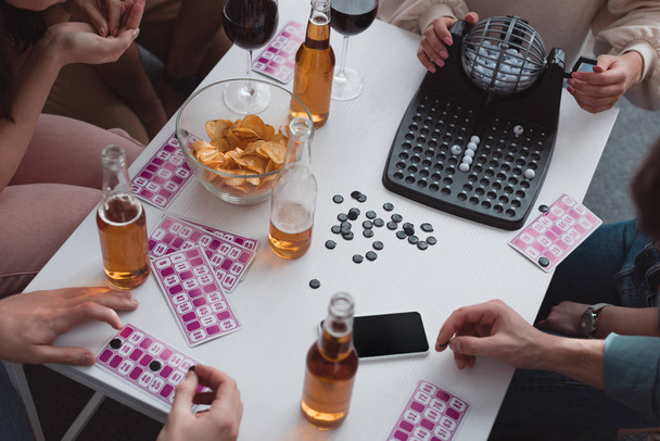 KYIV, UKRAINE - JANUARY 27, 2020: cropped view of friends sitting at table with lotto balls dispenser, cards, covering chips and smartphone with blank screen - Photo, Image