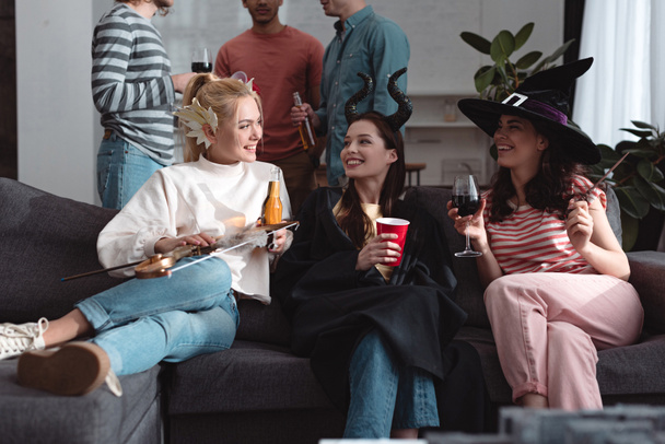 cropped view of men standing near cheerful girls in fairy costumes drinking beverages on sofa - Фото, изображение