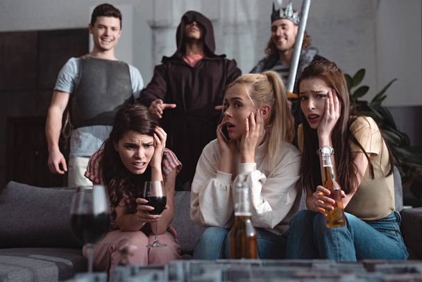 σοκαρισμένα κορίτσια πίνοντας μπύρα και κρασί, ενώ οι άνδρες με κοστούμια νεράιδες χαρακτήρες στέκεται στο παρασκήνιο - Φωτογραφία, εικόνα