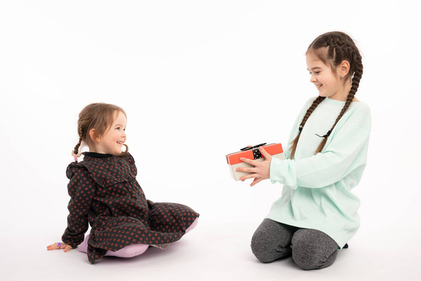 Belle adolescente en sweat à capuche bleu montre boîte cadeau à sa petite soeur rousse en robe, isolé sur fond blanc
 - Photo, image