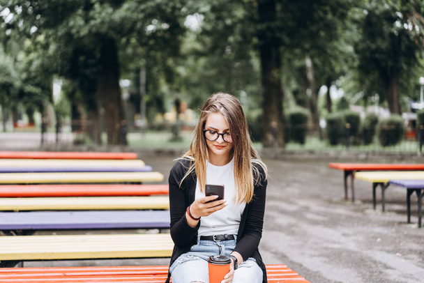 Young girl in glasses looks into a cellphone. In her second hand holds a cup of coffee sitting on a park bench. Concept of communication and social networks. - Фото, изображение