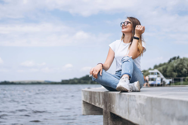 Young woman with long hair in stylish glasses posing on the concrete shore near the lake. Girl dressed in jeans and t-shirt smiling and looking away. - Photo, Image