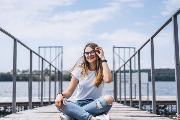 Young woman with long hair in stylish glasses posing on a wooden pier near the lake. Girl dressed in jeans and t-shirt smiling and looking at the camera. - Photo, image