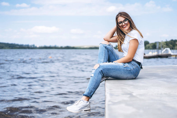 Young woman with long hair in stylish glasses posing on the concrete shore near the lake. Girl dressed in jeans and t-shirt smiling and looking at the camera. - Fotó, kép