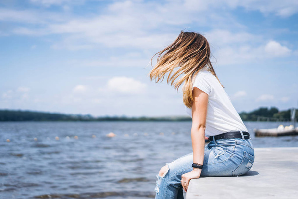 Young woman with long hair in stylish glasses posing on the concrete shore near the lake. Girl dressed in jeans and t-shirt smiling and looking away. - Φωτογραφία, εικόνα