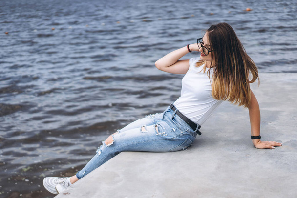 Young woman with long hair in stylish glasses posing on the concrete shore near the lake. Girl dressed in jeans and t-shirt smiling and looking away. - Zdjęcie, obraz