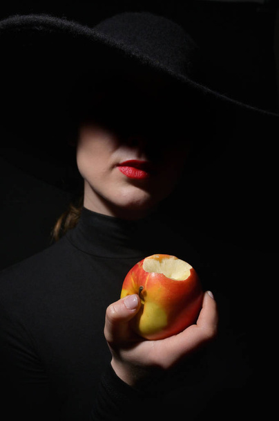 Μια γυναίκα με κόκκινα χείλη σε ένα μαύρο καπέλο κρατώντας δαγκωμένο μήλο - Φωτογραφία, εικόνα